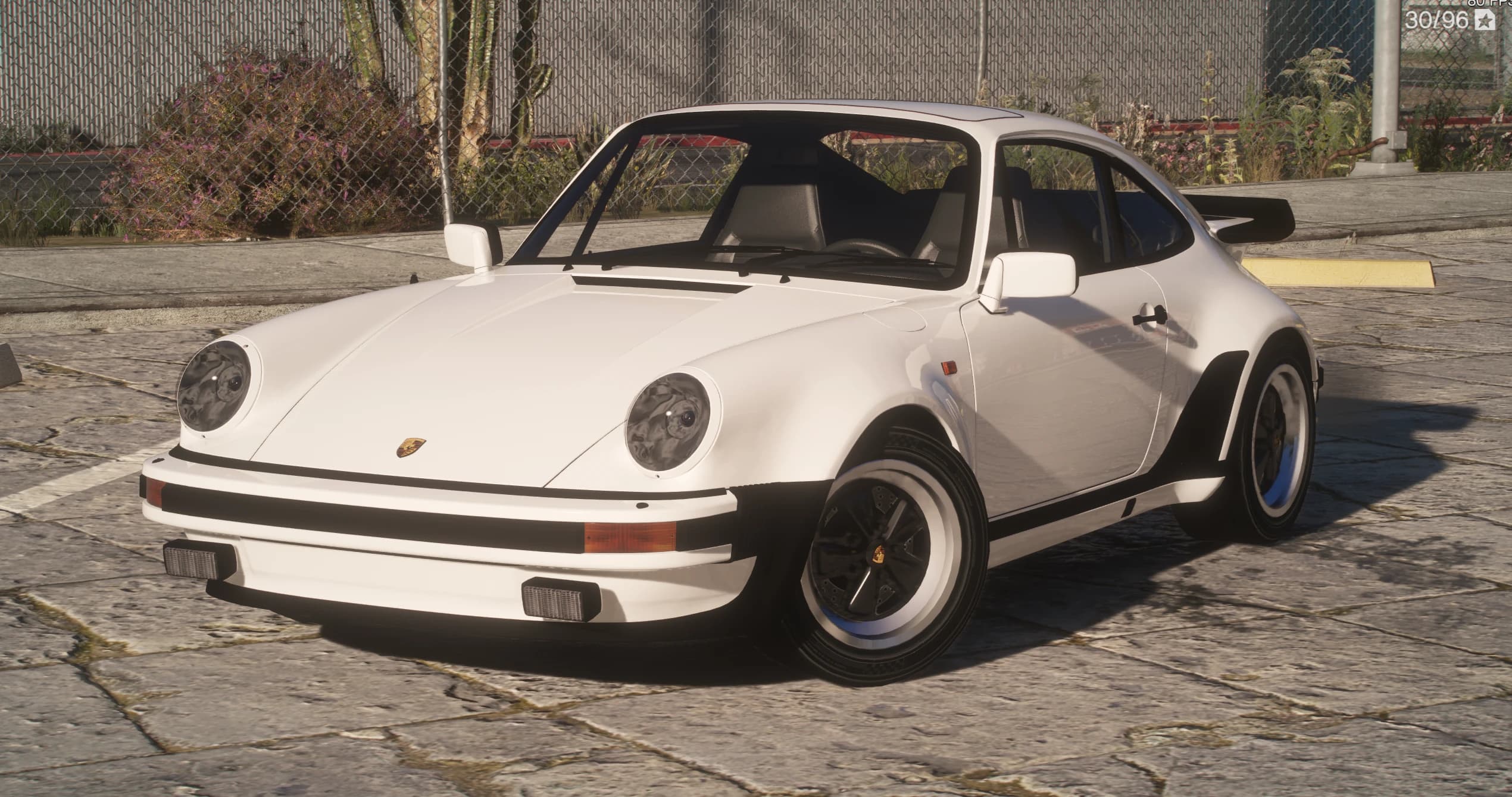 Porsche 911 Turbo 3.3 1982 en blanco, vista frontal en tres cuartos sobre losas de piedra, con los faros redondos, los paragolpes de banda de goma negra, el escudo de Porsche en el capó y las llantas escalonadas de estilo Fuchs bajo aletas ensanchadas