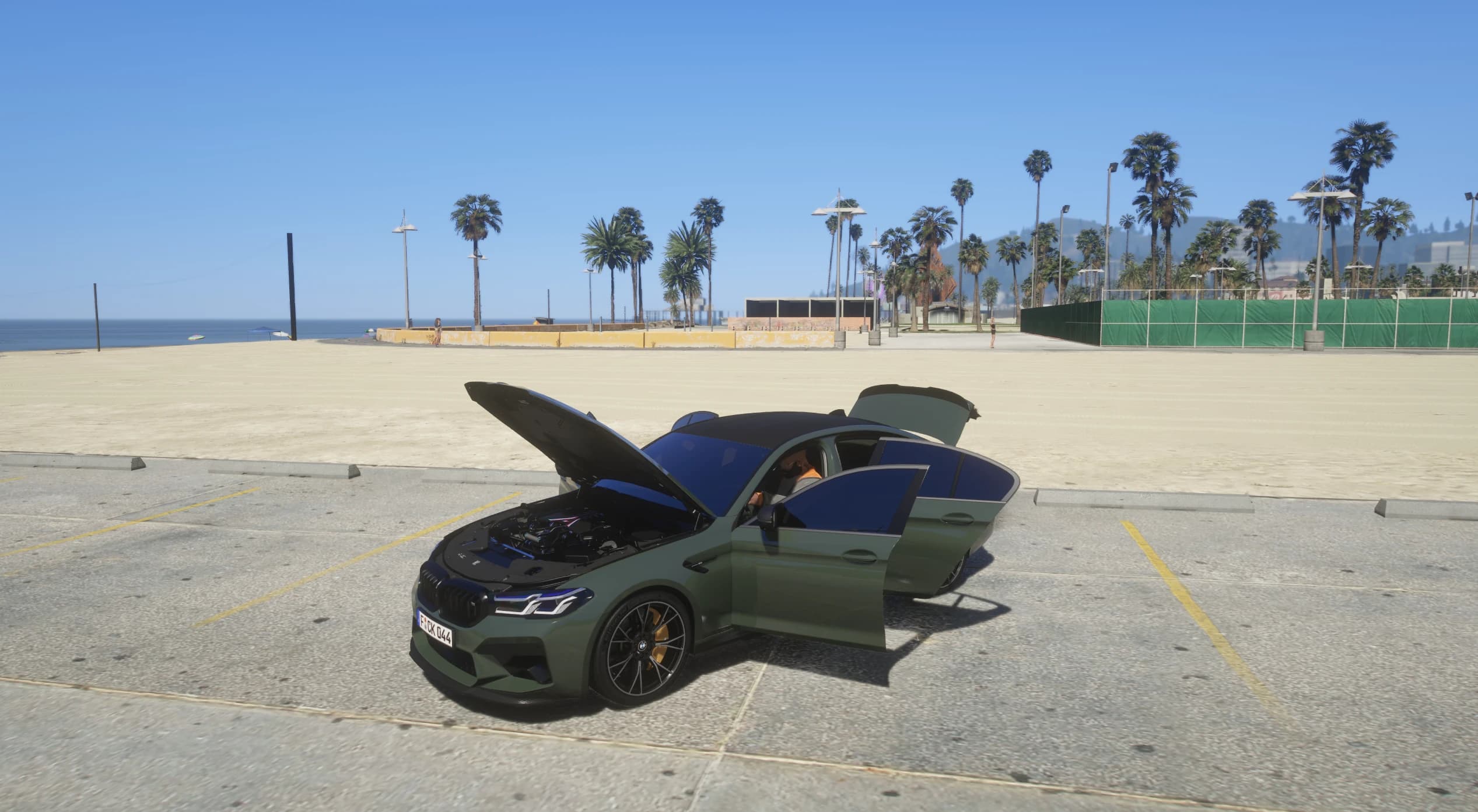 El M5 con el capó y la puerta del conductor abiertos, dejando ver el vano motor con detalles azules y un personaje sentado dentro, aparcado junto a la playa con palmeras y el mar al fondo