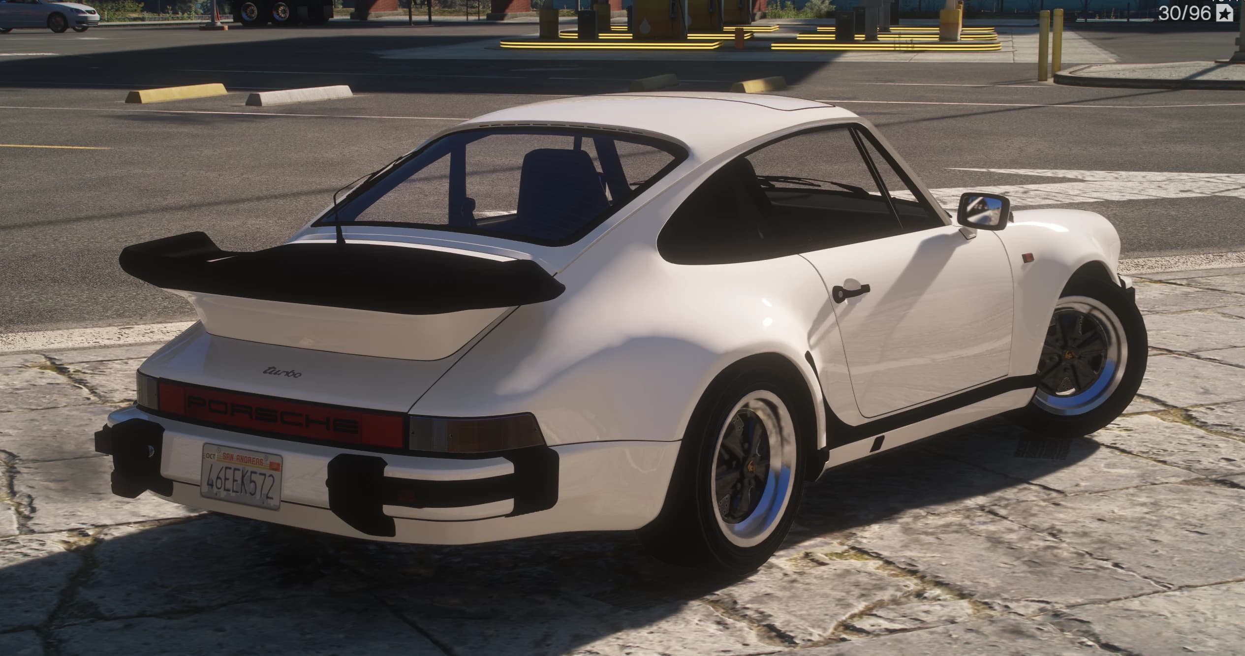 Tres cuartos trasero del 911 Turbo blanco en una gasolinera, con el alerón cola de ballena de labio negro, el rótulo 'turbo' en la tapa del motor, la banda reflectante roja 'PORSCHE' y una matrícula de San Andreas con el texto 46EEK572