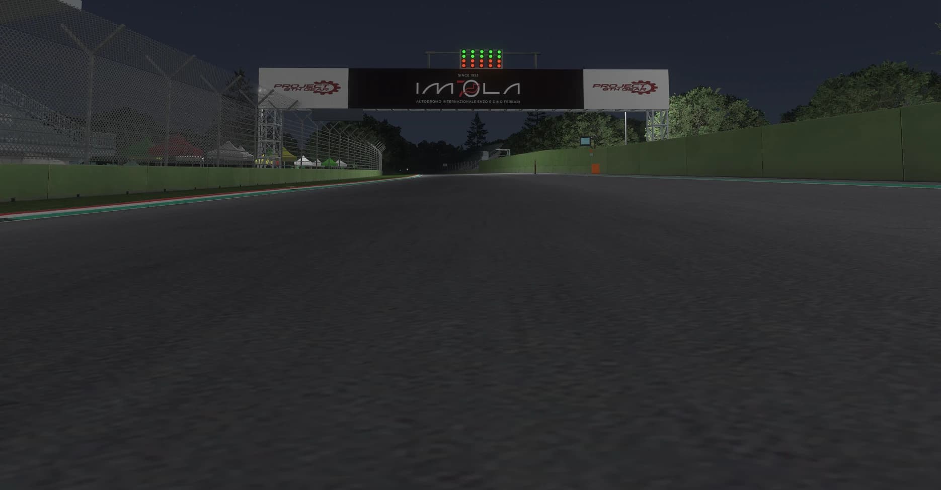 Vista nocturna desde la línea de salida de Imola mirando recta arriba: el pórtico iluminado de IMOLA con el rótulo Autodromo Internazionale Enzo e Dino Ferrari, el semáforo de salida encima, la tribuna a la izquierda y los muros verdes del trazado