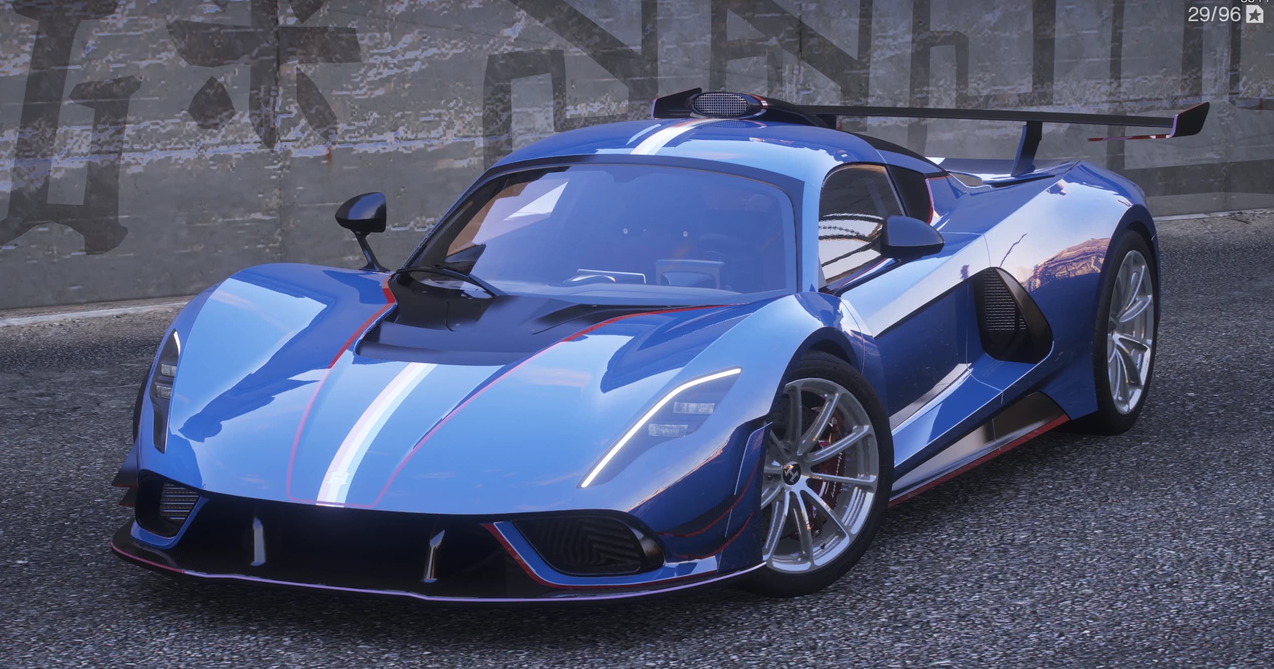 Hipercoche Hennessey Venom F5 Revolution Roadster azul con franja central blanca y roja y un gran alerón trasero, tres cuartos frontal ante un muro con grafiti