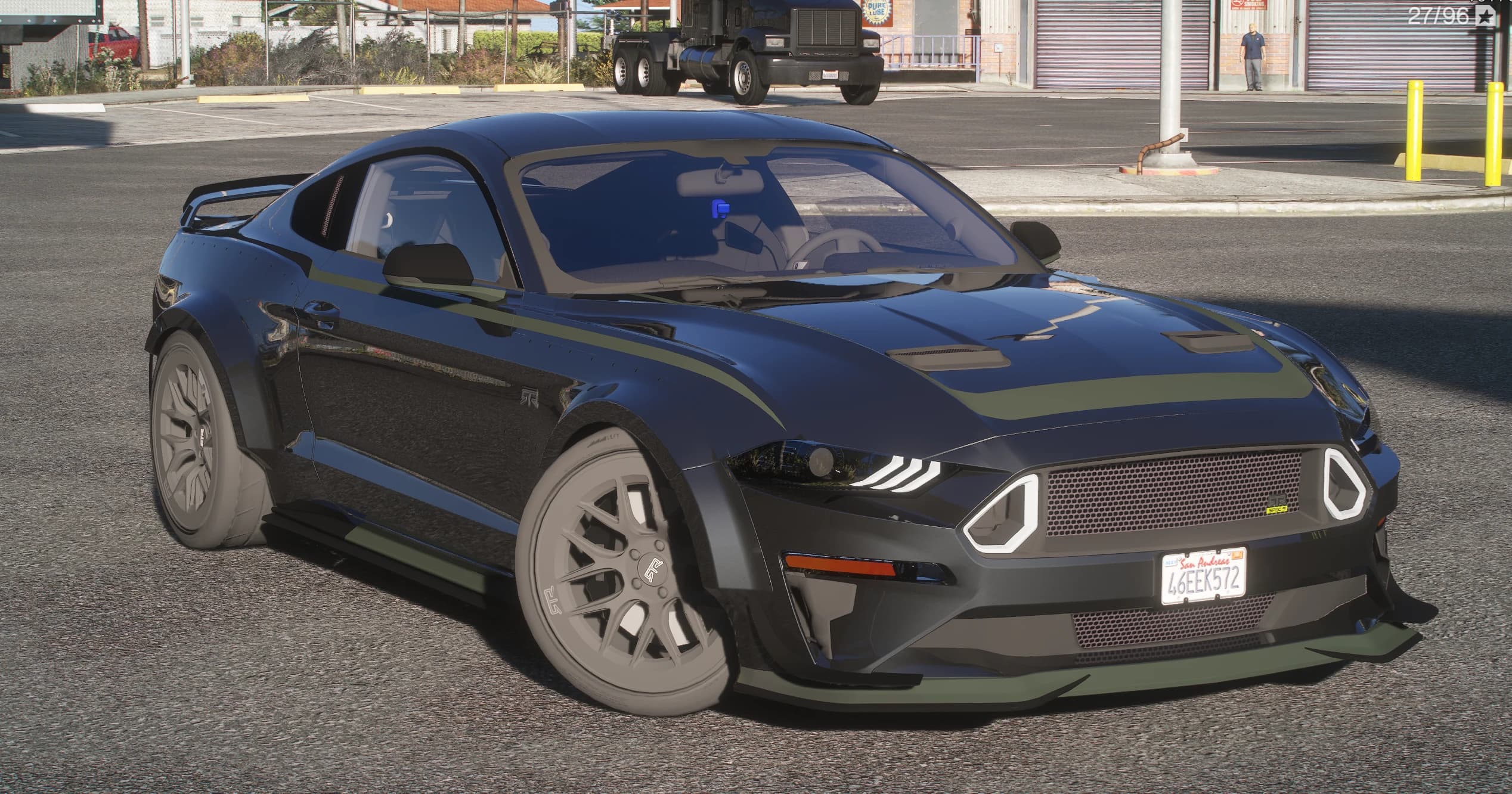 Ford Mustang RTR Spec 5 2015 en negro en una explanada industrial, vista frontal en tres cuartos con franjas verde oliva sobre el capó, aletas ensanchadas atornilladas, un splitter delantero profundo y llantas multirradio grises