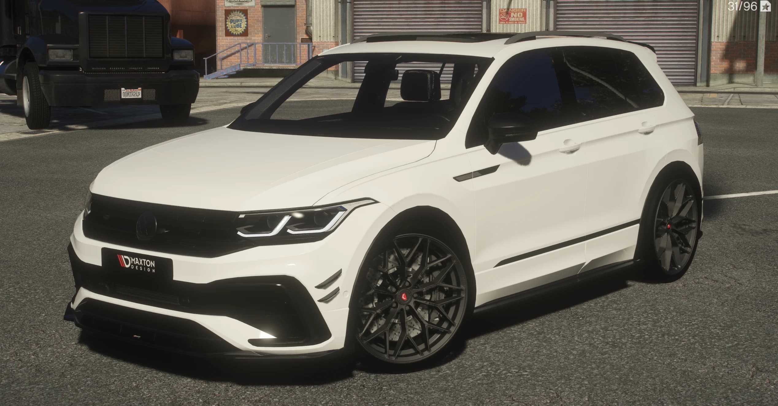 Vista frontal en tres cuartos del Tiguan R blanco con la placa de Maxton Design en el splitter y llantas oscuras de radios múltiples con discos perforados, aparcado junto a un camión negro