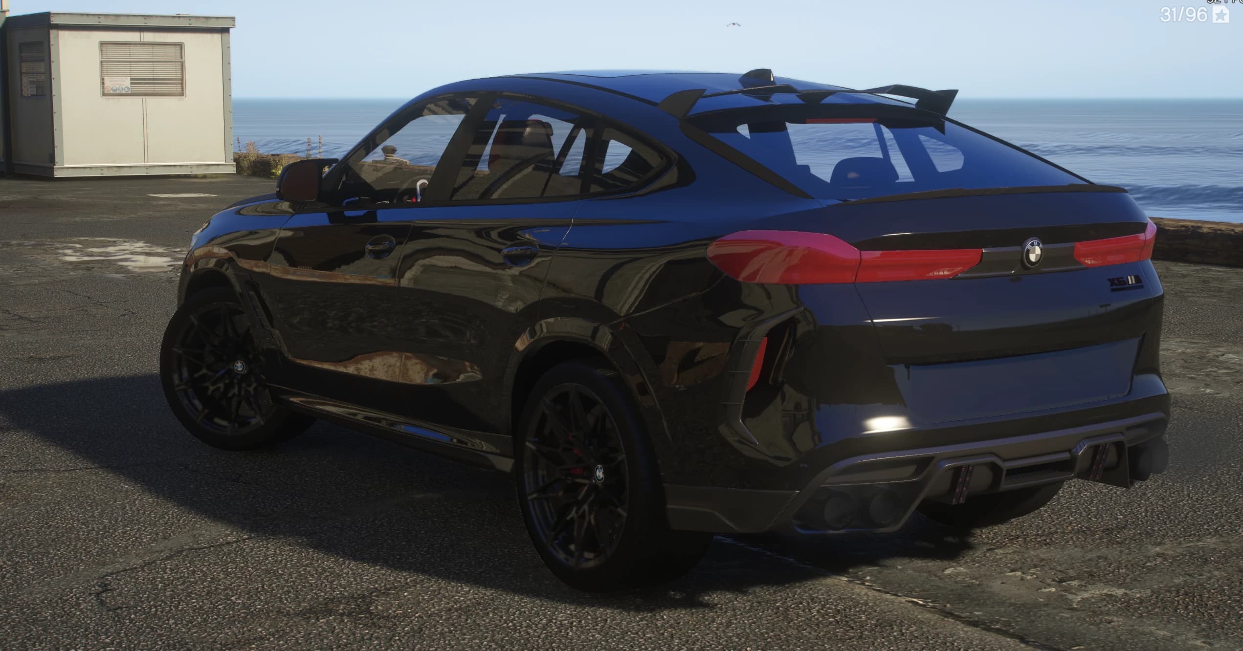 Vista trasera en tres cuartos junto al mar con el alerón de techo con aletas laterales, la insignia X6 M, las cuatro salidas de escape y llantas negras de radios