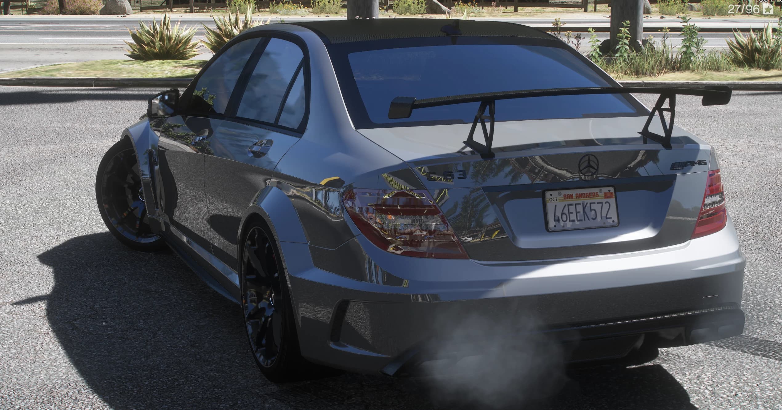 Tres cuartos trasero del C63 plata en la cuneta, con el gran alerón negro sobre sus soportes, los emblemas C 63 y AMG, el techo de carbono y humo saliendo de la cuádruple salida de escape