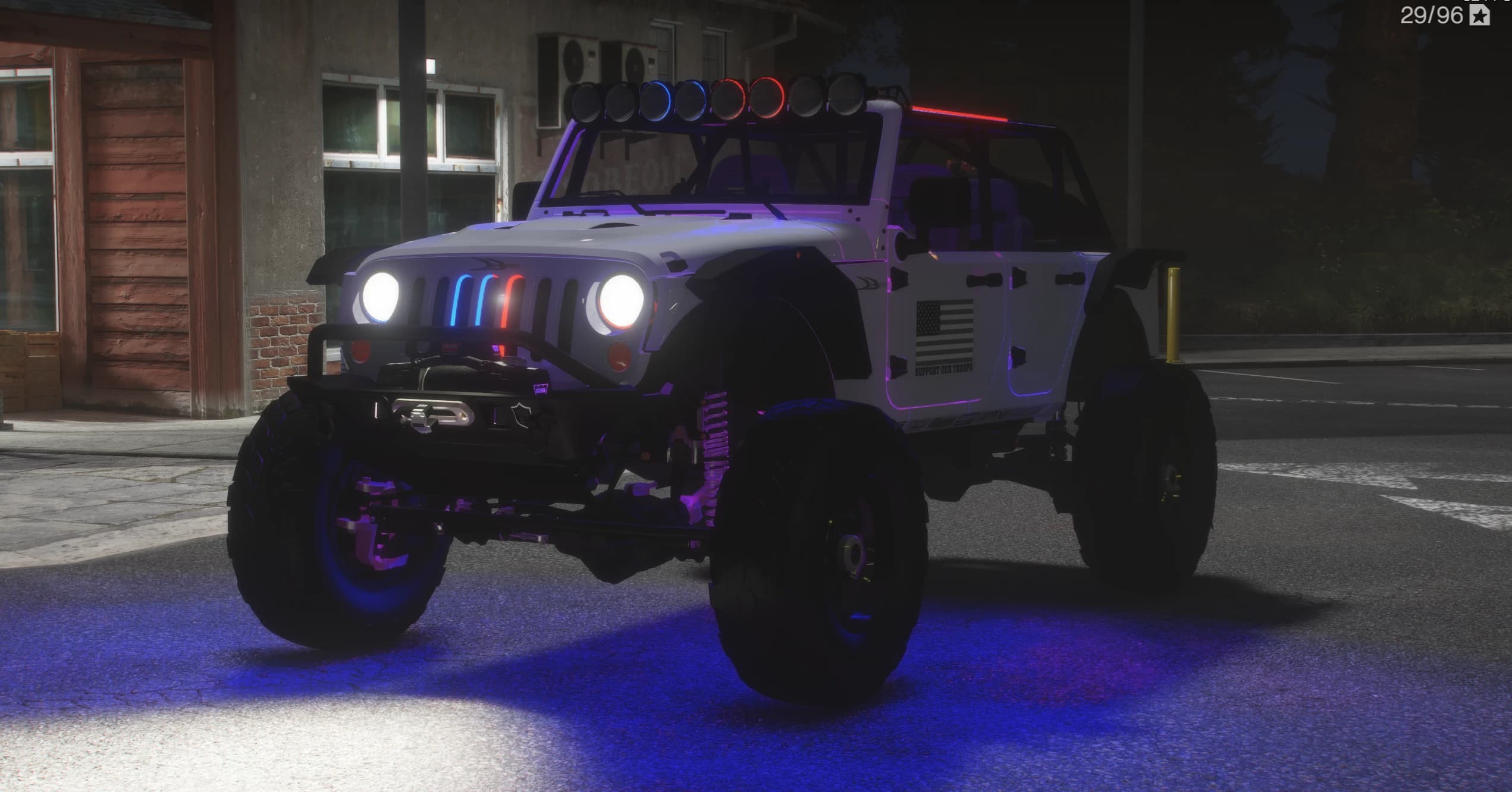 Jeep Wrangler blanco preparado para el campo y elevado, de noche, con barra de focos en el techo, jaula, parachoques con cabrestante, neumáticos de barro sobredimensionados y luz inferior azul, con calcas de bandera de EE. UU. y Support Our Troops en las puertas