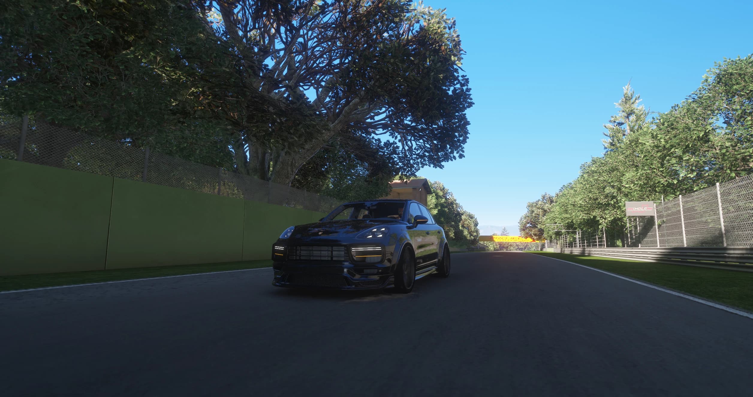 El Cayenne rodando por un tramo arbolado del circuito de Imola por la mañana con las luces diurnas LED y los intermitentes delanteros ámbar encendidos, y un cartel de IMOLA en la valla de la derecha