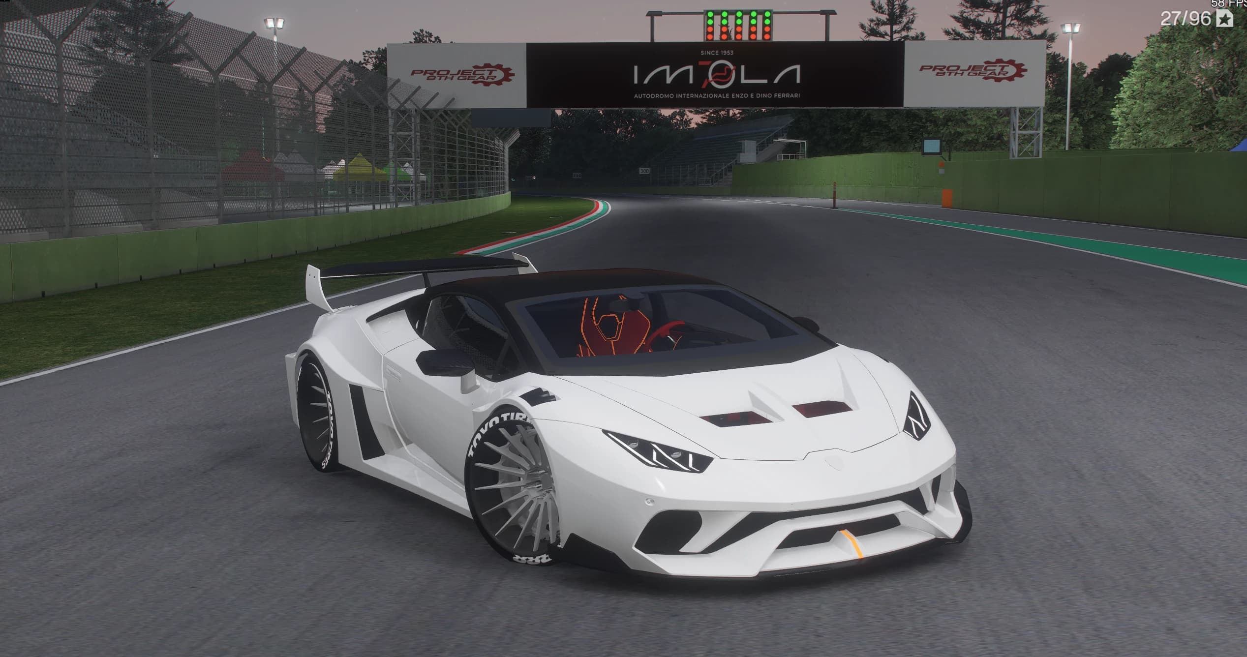 Lamborghini Huracán blanco con el ancho de vía Liberty Walk aparcado en la recta de salida de Imola al anochecer, en tres cuartos delantero bajo el pórtico de IMOLA, con los pasos de rueda atornillados, la jaula antivuelco tras el cristal y el alerón de cuello de cisne