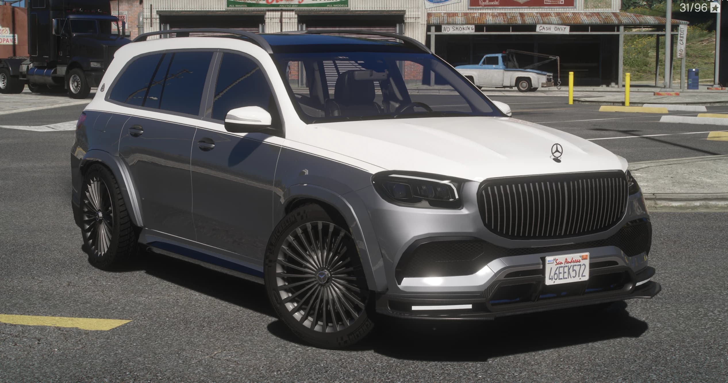 Vista frontal en tres cuartos del Maybach GLS600 bitono blanco sobre gris aparcado ante un taller viejo, con la parrilla de lamas verticales, el splitter delantero de carbono y llantas de estilo turbina
