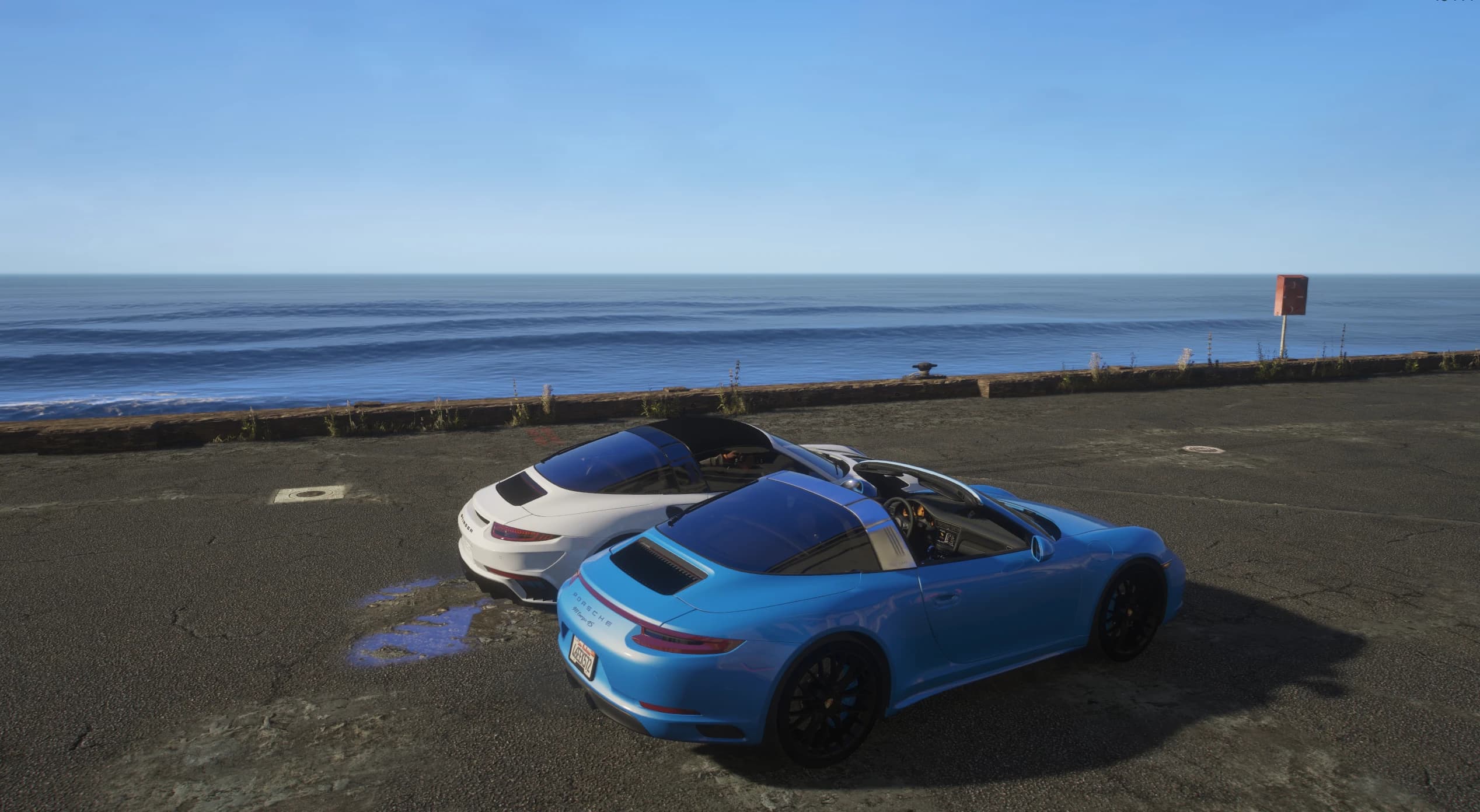 Tres cuartos trasero en picado de ambos coches aparcados junto al murete del mar, con el Targa 4S azul y su arco Targa plateado en primer plano y el Stinger blanco detrás