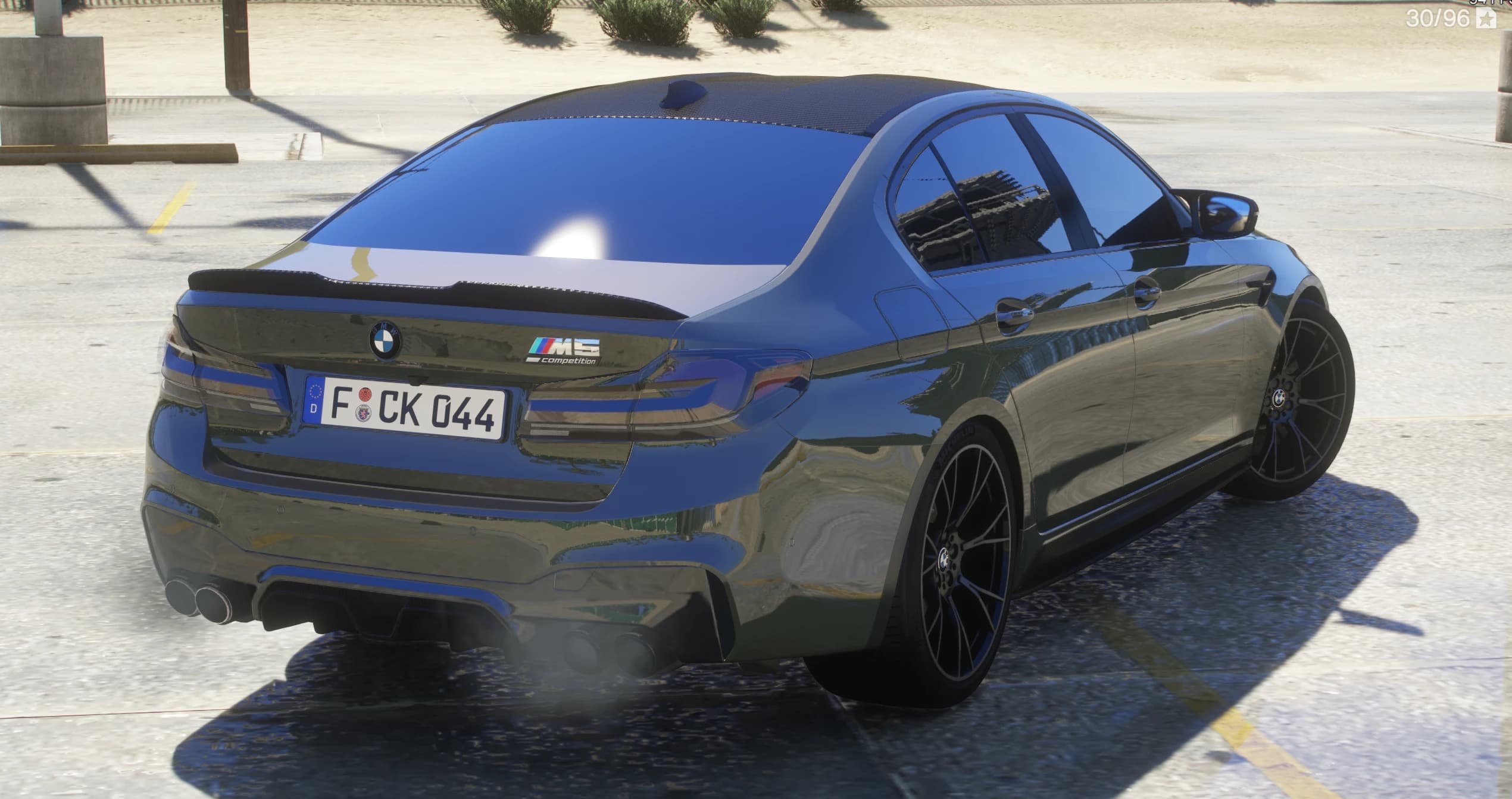 Tres cuartos trasero del M5 oliva con techo de fibra de carbono, alerón ducktail con el emblema M5 Competition, los cuatro escapes soltando humo y llantas negras multirradio, en un aparcamiento soleado