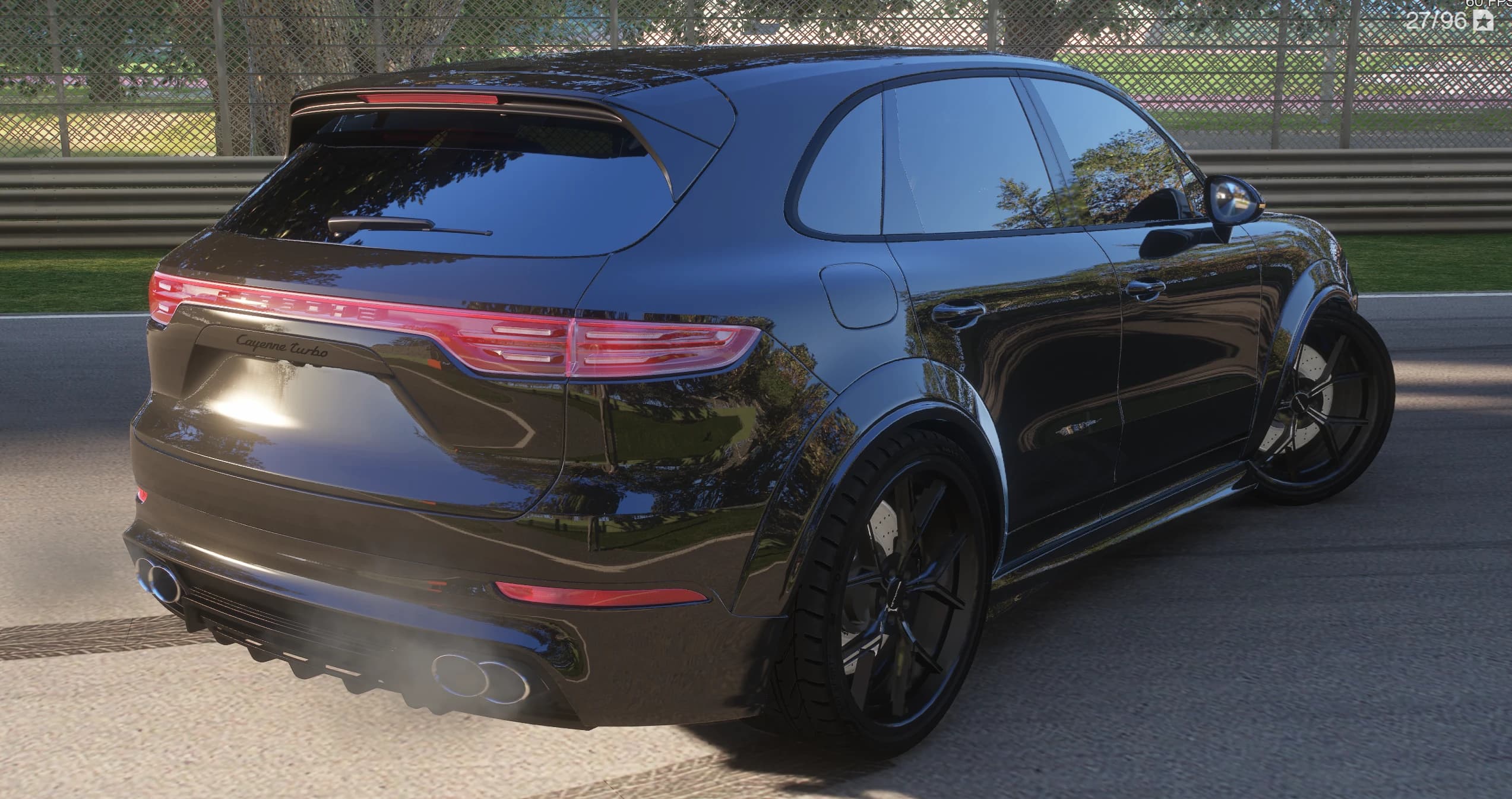 Tres cuartos trasero del Cayenne negro con la barra de luces Porsche encendida a lo ancho del portón, el rótulo Cayenne Turbo, el alerón de techo y los escapes soltando humo junto a la barrera del circuito