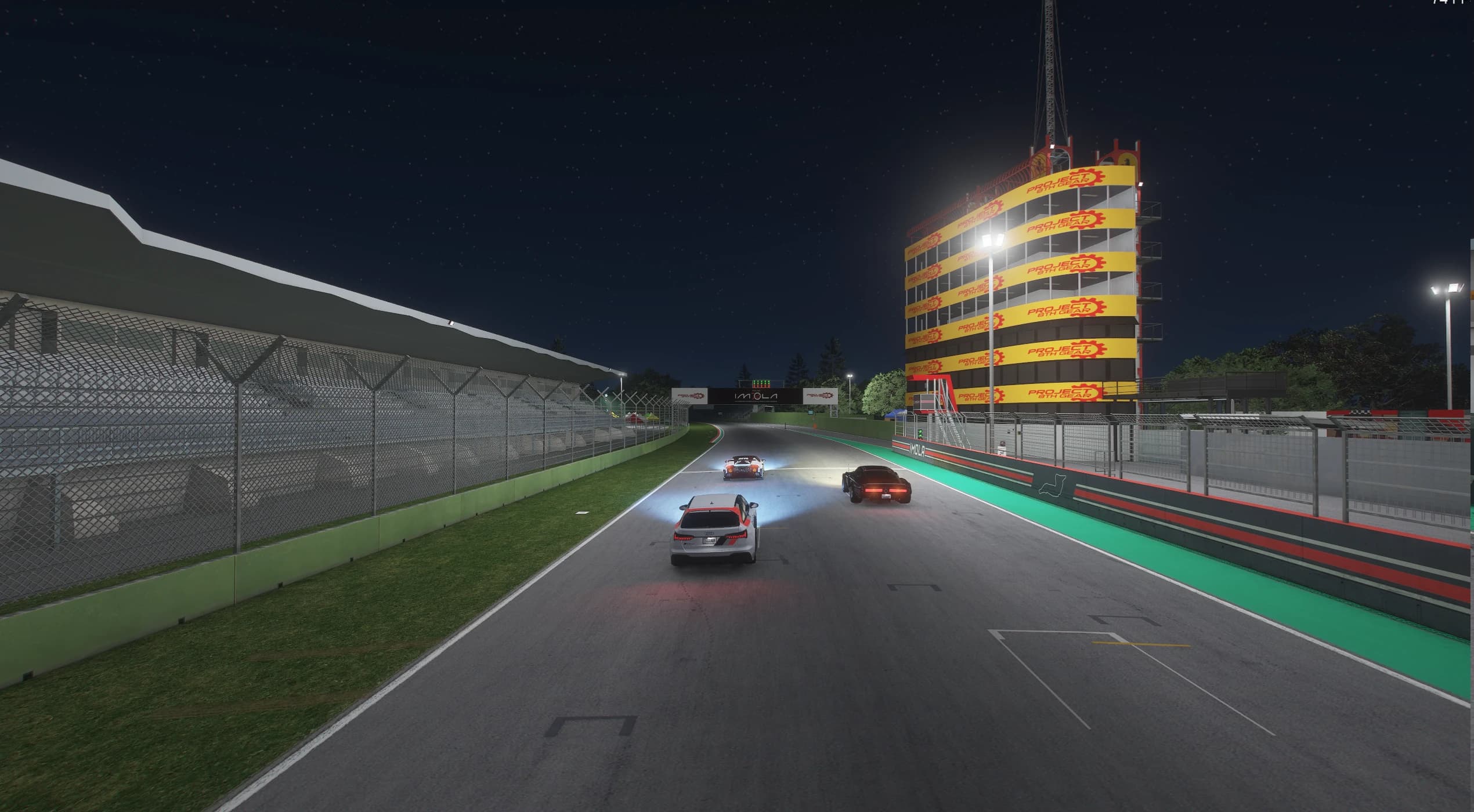 Tres coches rodando de noche por la recta de boxes iluminada, con la tribuna cubierta a la izquierda y la torre de boxes forrada de carteles de PROJECT 8TH GEAR a la derecha