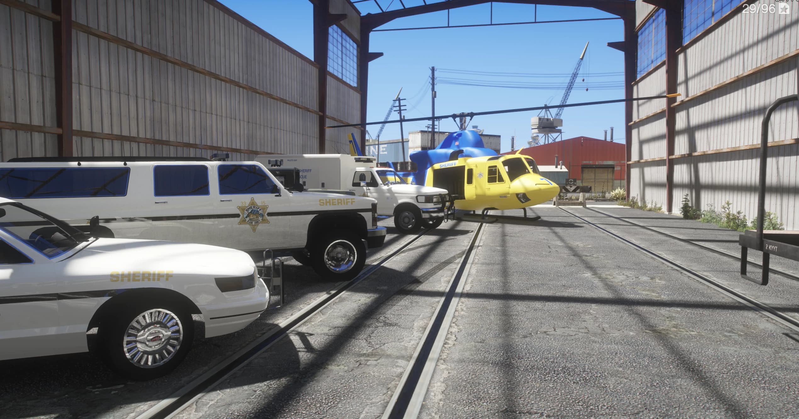 Vista a ras de suelo de la flota del sheriff alineada en un cobertizo: un coche patrulla, un todocamino del sheriff, un camión Paleto Bay Sheriff Rescue y un helicóptero del sheriff amarillo, todos con la librea retro