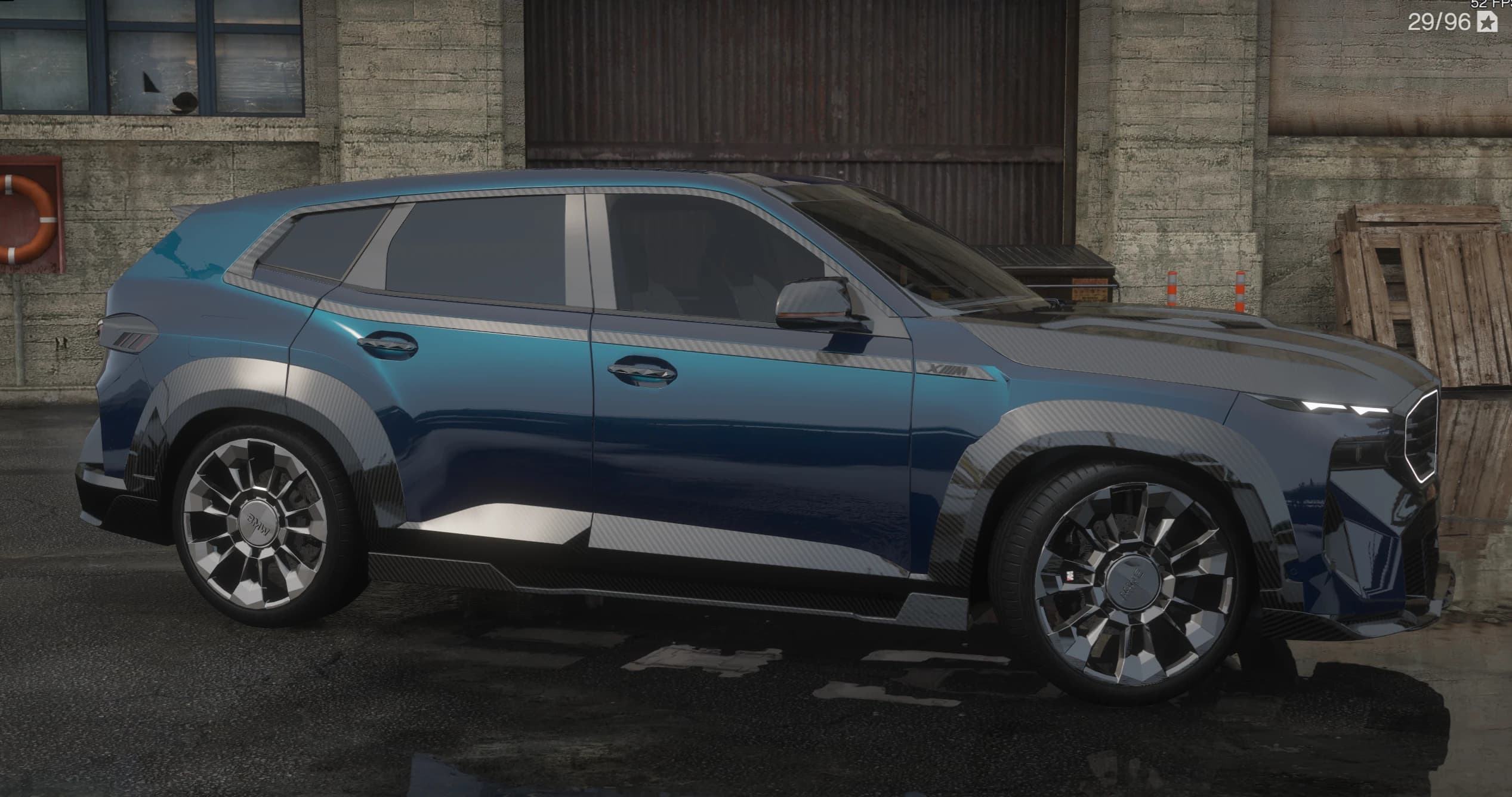 Perfil lateral del BMW XM azul con la parte baja de la carrocería en carbono, insignia XM y grandes llantas de varios radios junto a un edificio industrial