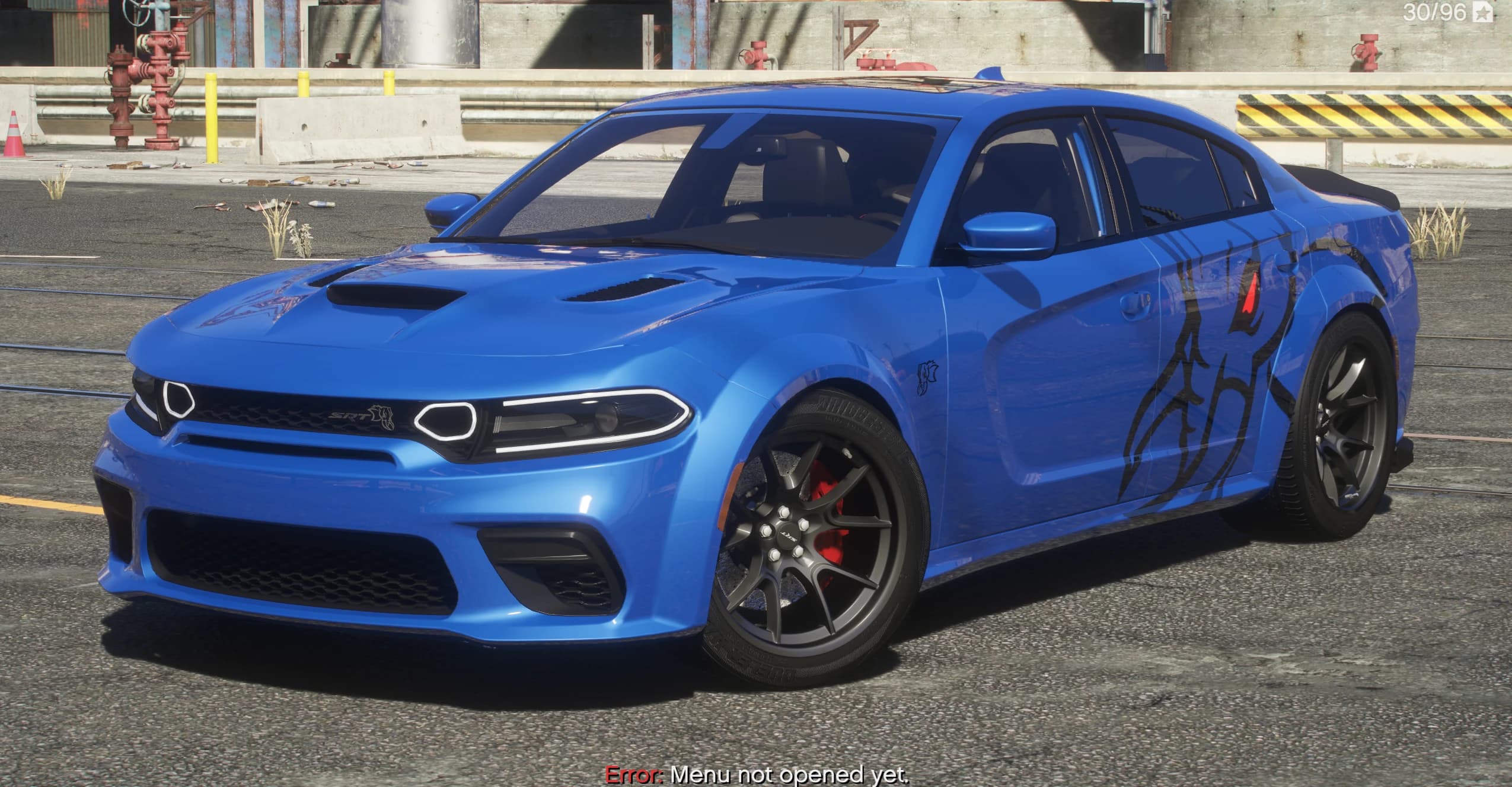 Dodge Charger Hellephant widebody 2024 en azul, vista frontal en tres cuartos en un patio de refinería industrial, con el emblema SRT de la parrilla, las dos tomas del capó, un gráfico tribal negro en el lateral y llantas grises con las pinzas de freno rojas