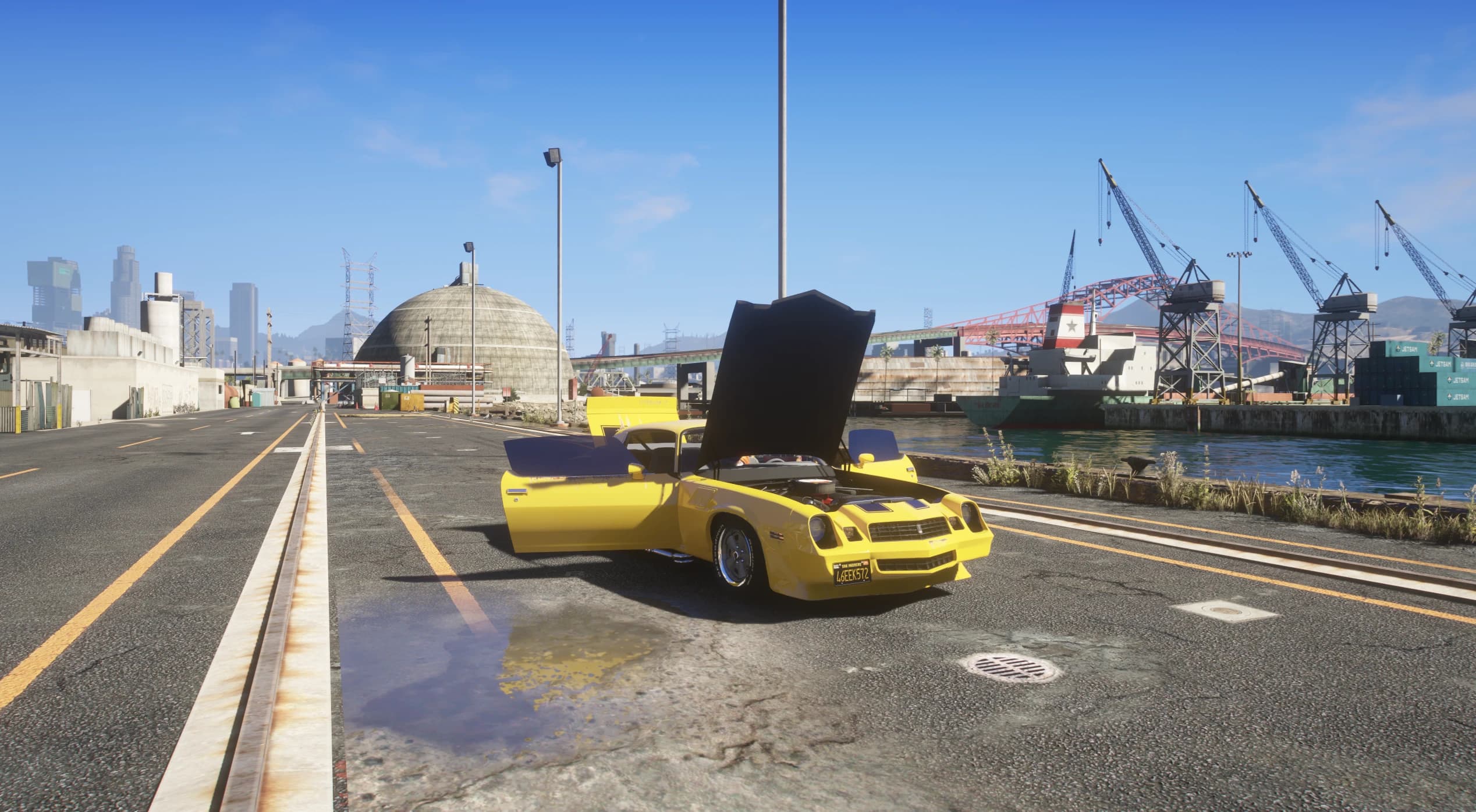 El Camaro con puertas y capó abiertos en una vía del puerto, con el motor a la vista y grúas y un carguero de fondo