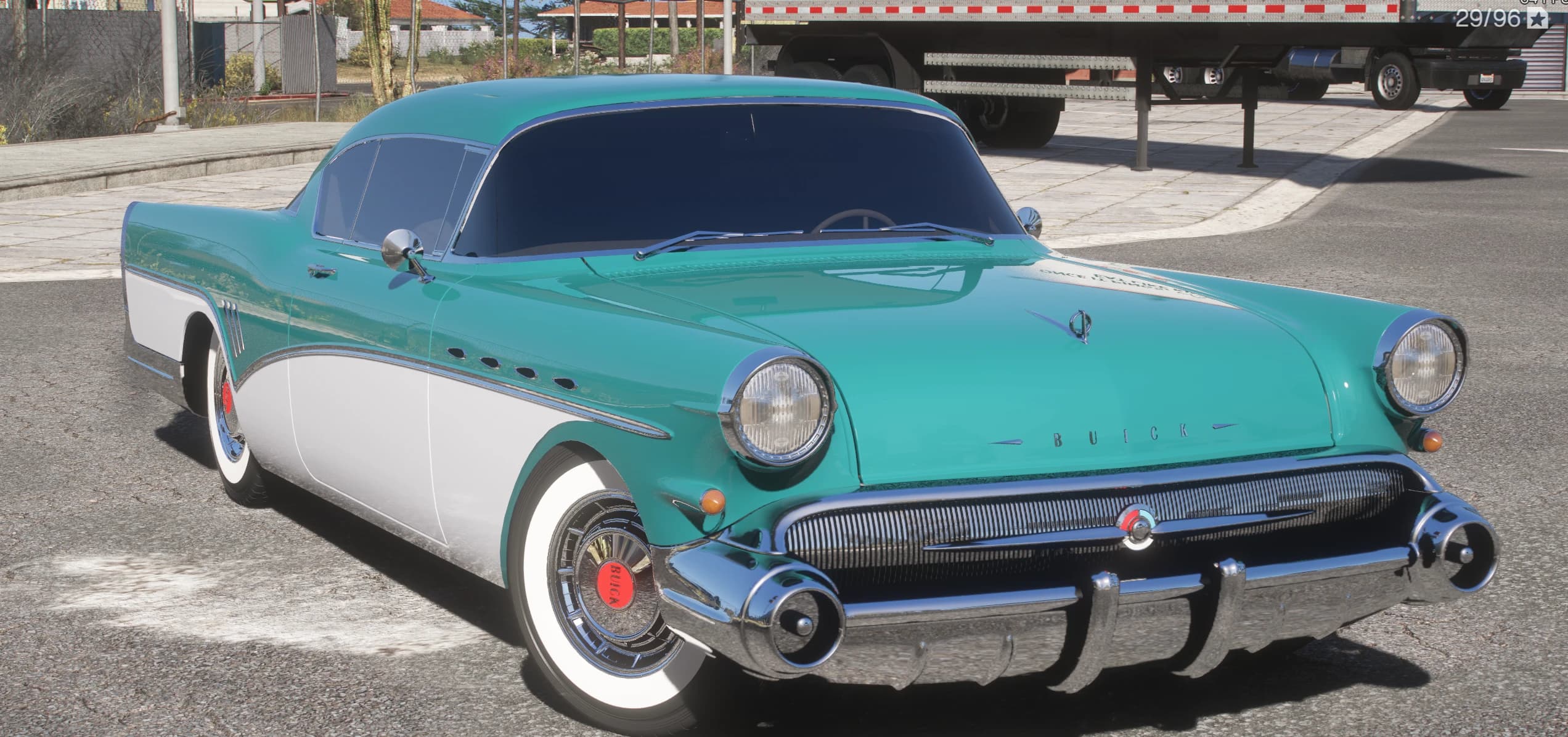 Buick Roadmaster de 1957 en verde azulado y blanco, tres cuartos frontal con parrilla cromada, faros con visera y neumáticos de banda blanca, aparcado en un área de camiones del desierto