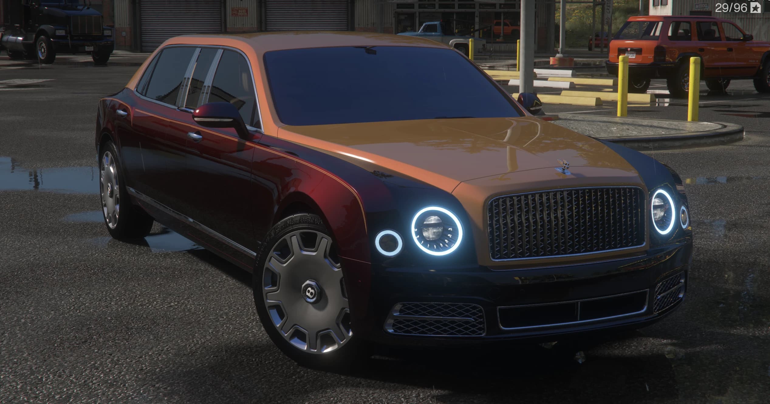 Limusina Bentley Mulsanne bitono en granate y cobre, tres cuartos frontal con faros de anillo iluminado, la insignia alada de Bentley en la parrilla y tapacubos con el logo B, en un área de camiones