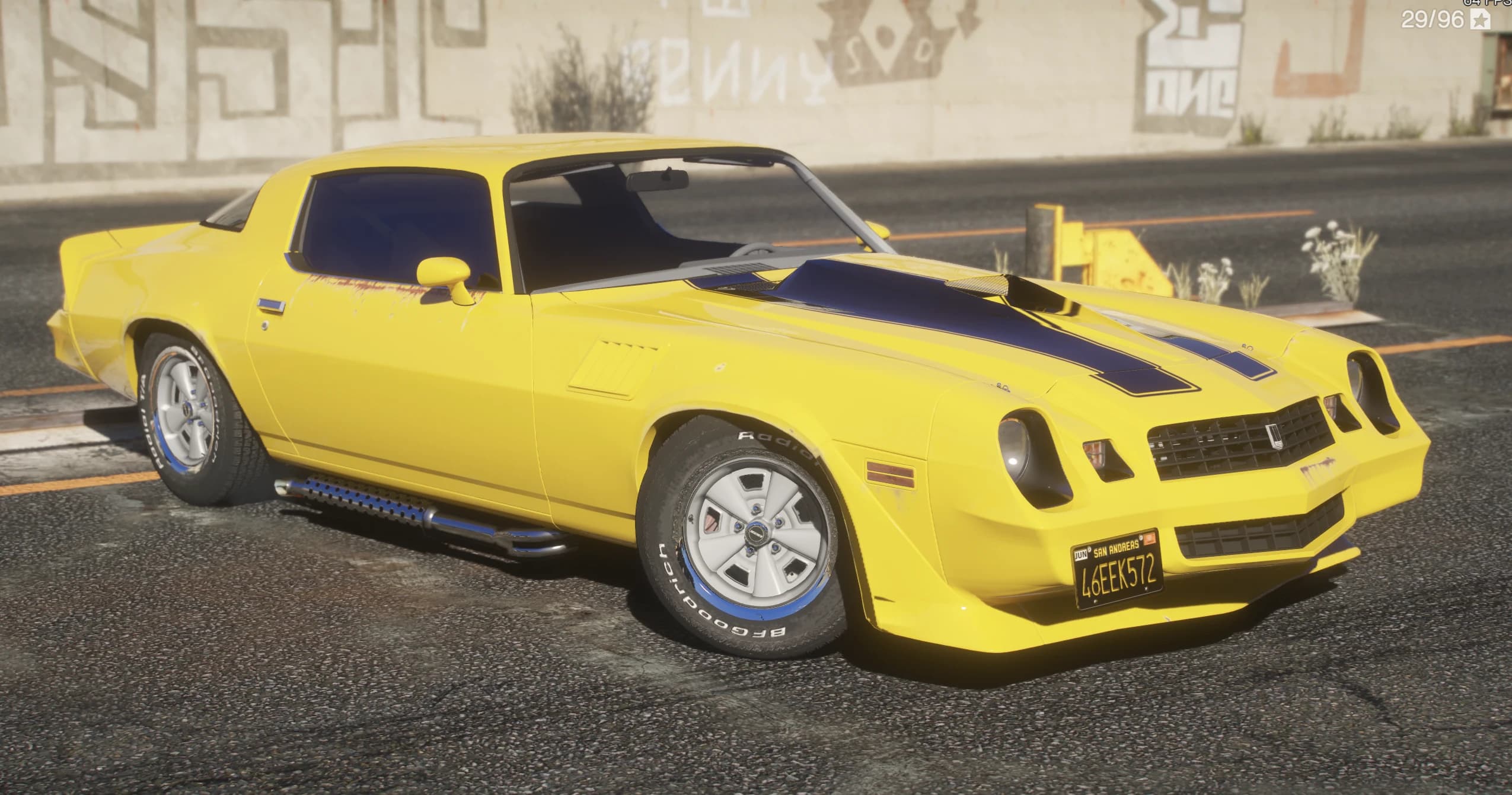 Vista frontal en tres cuartos del Camaro Z28 amarillo con franjas negras en el capó y neumáticos BFGoodrich de letras blancas junto a un muro con grafitis
