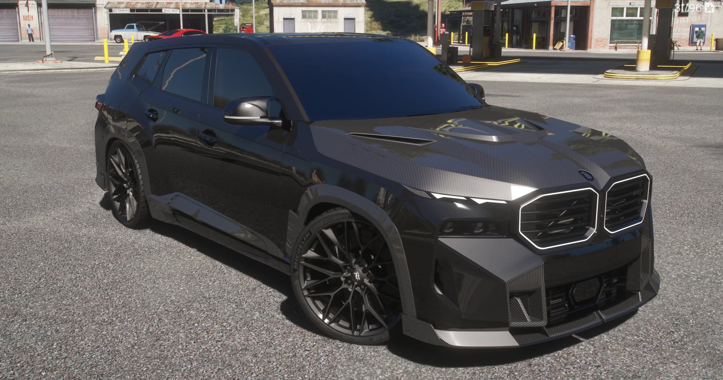 Vista frontal en tres cuartos del BMW XM Larte negro en una gasolinera rural, con el capó de carbono, el contorno iluminado de las parrillas y neumáticos Michelin