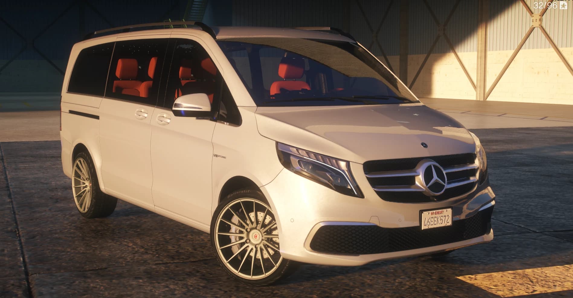 Vista frontal en tres cuartos del V-Class blanco en un hangar con la parrilla estilo AMG, el emblema V8 Biturbo en la aleta, llantas de radios de turbina y asientos rojos dentro