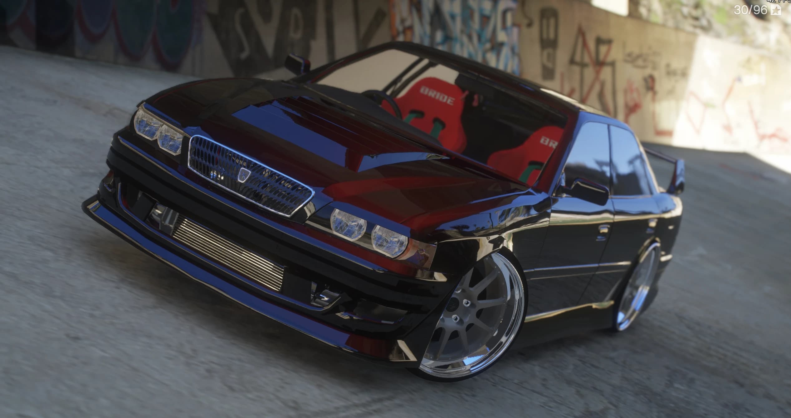 Vista frontal en tres cuartos del Toyota Chaser JZX100 negro bajo un paso elevado lleno de grafitis, con el intercooler frontal a la vista, los faros dobles y los baquets rojos estilo BRIDE tras el parabrisas