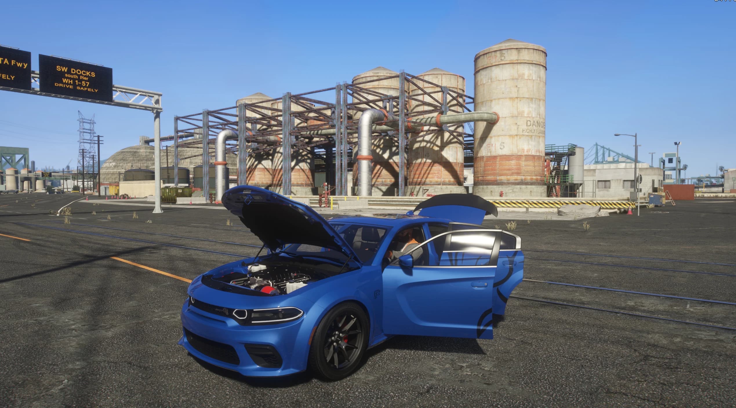 Charger azul con el capó y la puerta del conductor abiertos en los muelles, dejando ver el vano motor HEMI sobrealimentado con la admisión roja, bajo un cartel de 'SW DOCKS' y grandes tanques de almacenamiento inflamable