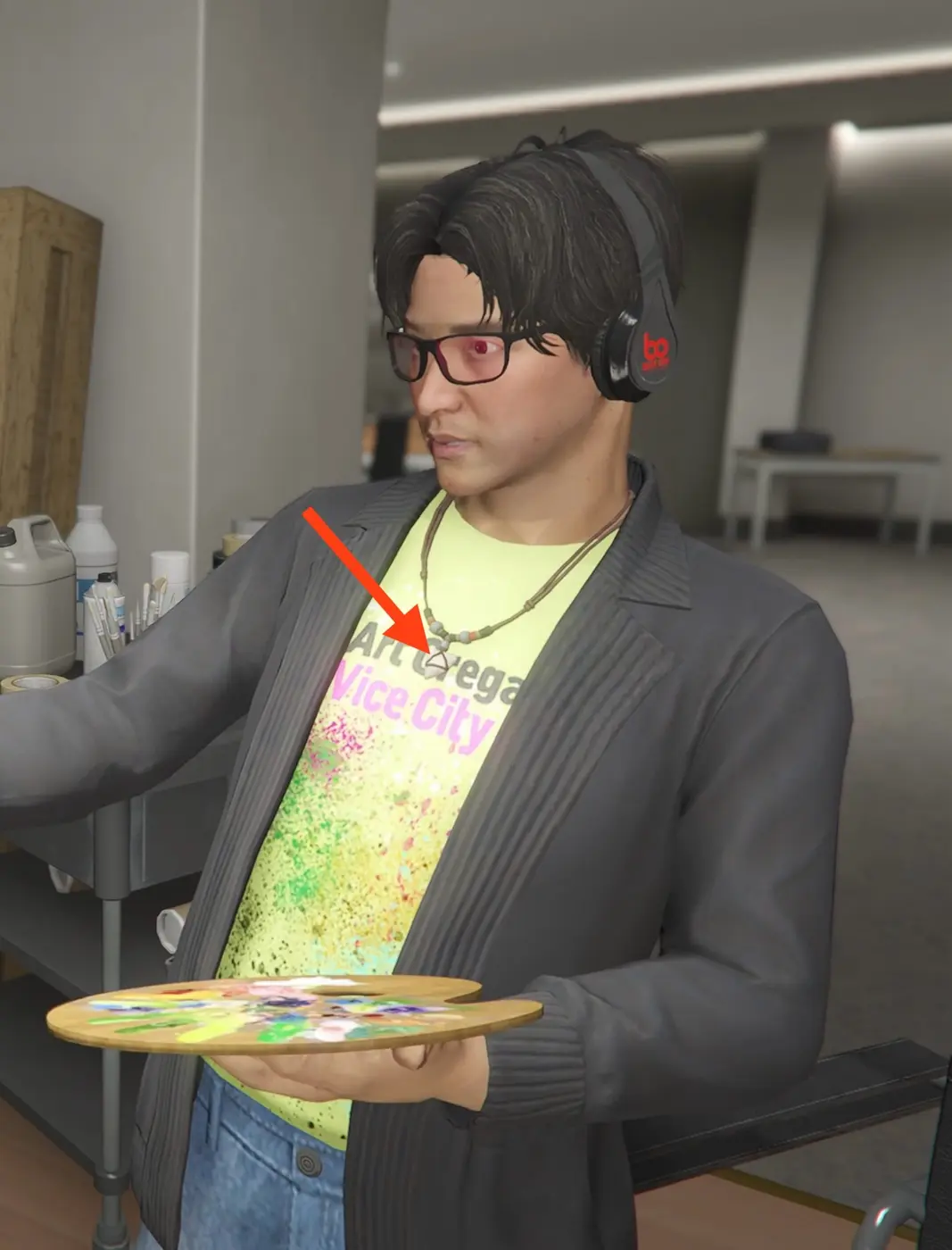 Un personaje de GTA Online con gafas y cascos sosteniendo una paleta de pintor, con una camiseta amarilla chillón que dice 'Art Gallery Vice City' en letras rosas y una flecha roja señalando un colgante con forma de reloj de arena en un collar de cuentas.